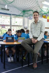 Teacher sitting on a desk with students in the classroom behind him